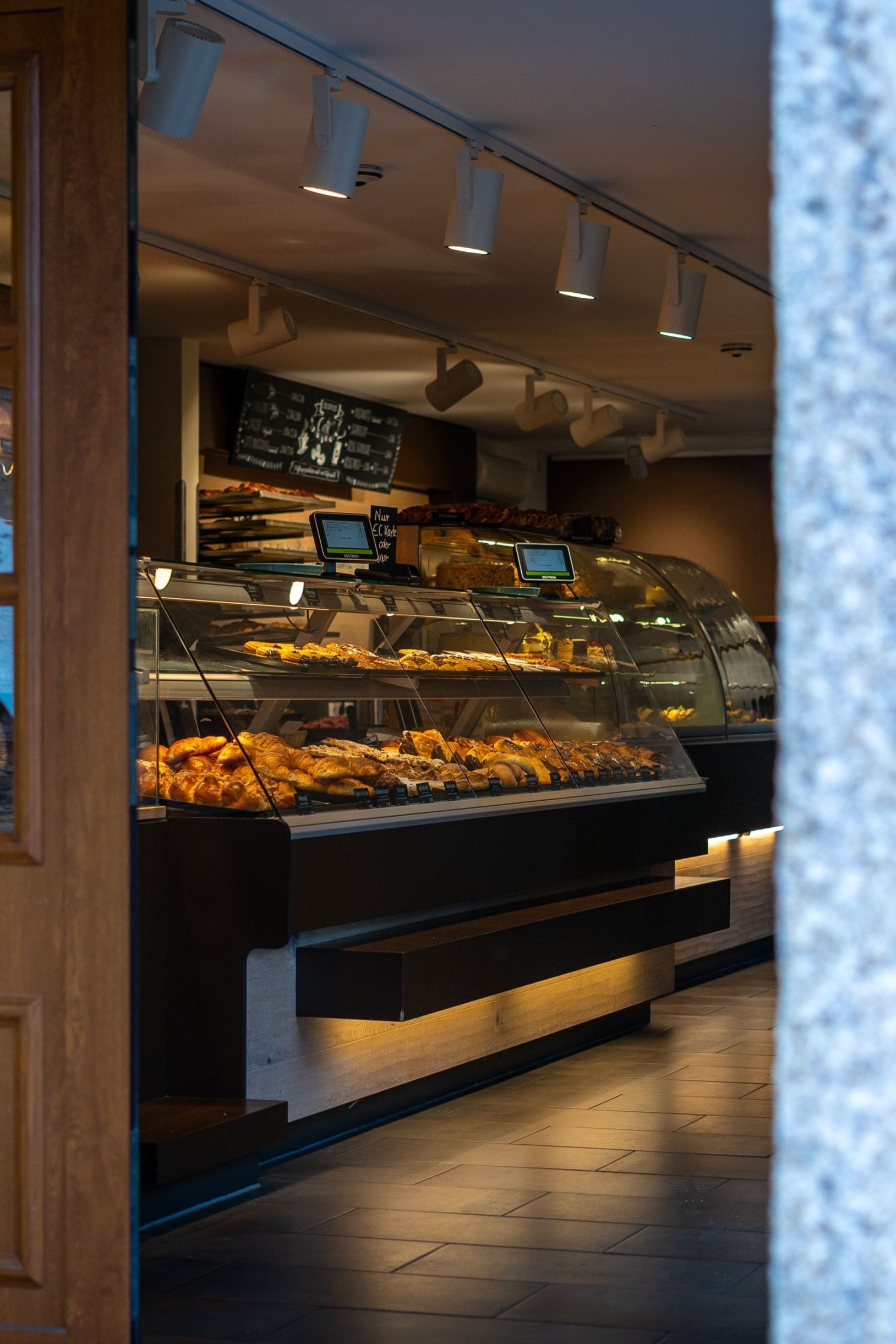 Ein Blick in eine Bäckerei mit einer Verkaufstheke, die verschiedene Backwaren wie Croissants, Brötchen und Kuchen zeigt, beleuchtet mit warmem Licht.