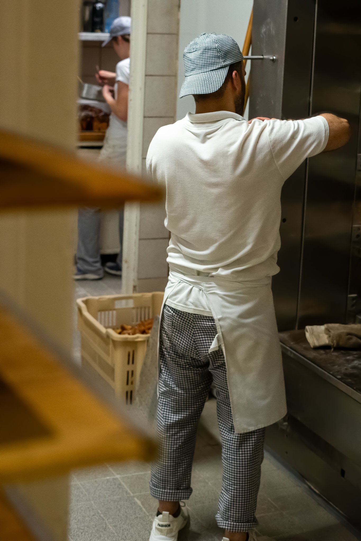 Bäcker vor Ofen in einer Backstube beim Arbeiten, trägt ein weißes T-Shirt, karierte Hosen und eine kariert Kappe.