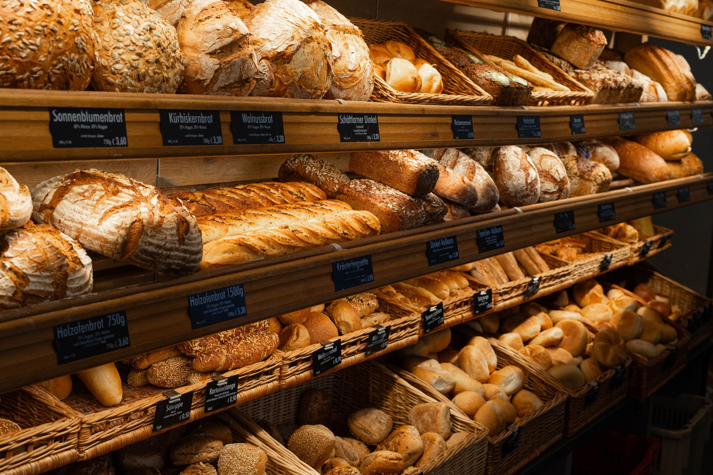 Verschiedene Sorten frisches Brot und Brötchen in einer Bäckerei, auf Holzregalen und in Körben ausgestellt.